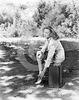 Young woman sitting on the side of the road on her suitcase, rubbing her aching feet