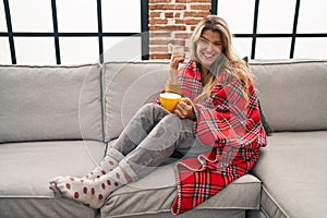 Young woman sitting on the sofa drinking a coffee at home doing happy thumbs up gesture with hand
