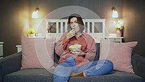 Young woman sitting on the sofa, eating popcorn and watching TV