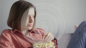 Young woman sitting on the sofa, eating popcorn and watching TV