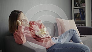 Young woman sitting on the sofa, eating popcorn, watching TV and falling alseep