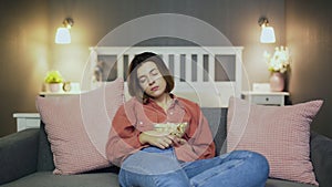 Young woman sitting on the sofa, eating popcorn, watching TV and falling alseep