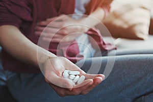 Young woman sitting on sofa with hand on aching stomach holding white medicine pills