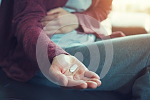 Young woman sitting on sofa with hand on aching stomach holding white medicine pills