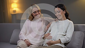 Young woman sitting on sofa with her pregnant friend, happy future mother