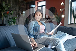 Young woman sitting on sofa, holding papers, typing on laptop