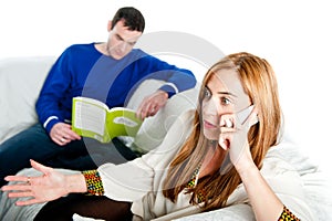 Young woman sitting on sofa at home, talking on a mobile whilst her boyfriend reads
