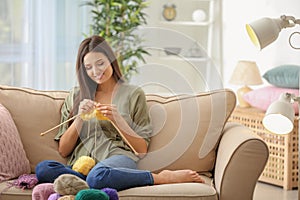 Young woman sitting on sofa and knitting warm sweater indoors