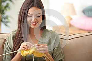 Young woman sitting on sofa and knitting warm sweater indoors