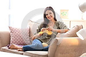 Young woman sitting on sofa and knitting warm sweater indoors