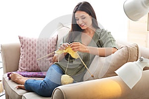 Young woman sitting on sofa and knitting warm sweater indoors