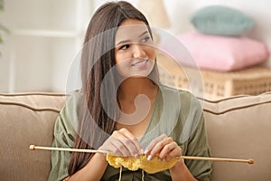 Young woman sitting on sofa and knitting warm sweater indoors