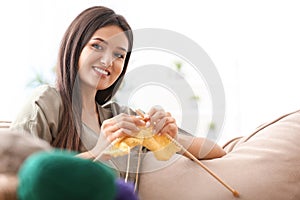 Young woman sitting on sofa and knitting warm sweater indoors