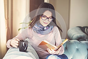 Young woman is sitting on a sofa and reading a book while holding a cup of coffee or tea