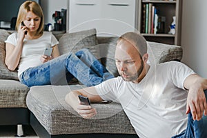 Young woman sitting at the sofa talking on phone while her husband is using his cellphone in their living room.