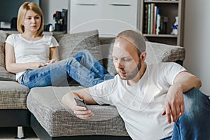 Young woman sitting at the sofa talking to her husband while he is using his cellphone in their living room.