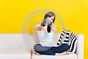 Young woman sitting on sofa wearing medical face mask protective show finger thumb up for good sign
