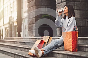 Young woman sitting on the stairs in the city, drinking coffee from a to go cup, with paper shopping bags around her