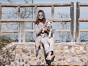 Young woman sitting on a stone fence with her Schauzer dog, poses and touches her hair looking straight ahead