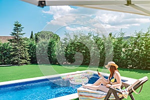 young woman sitting on a sun lounger in a bikini, by the pool looking at her smartphone. girl enjoying her summer