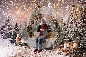 Young woman sitting on a swing with a blanket under the flashlights and drinkig a cup of coffee in a snow-covered park, concept