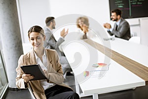 Young woman sitting by the table with digital tablet and paper chart in modern office in front of her team