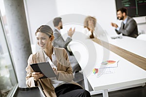 Young woman sitting by the table with digital tablet and paper chart in modern office in front of her team