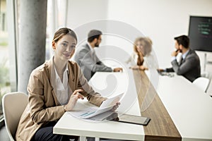 Young woman sitting by the table with digital tablet and paper chart in modern office in front of her team