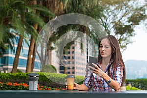 Young woman sitting at a table outdoors using her mobile phone. Female reading text messages on smartphone in the park.