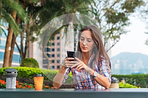 Young woman sitting at a table outdoors using her mobile phone. Female reading text messages on smartphone in the park.