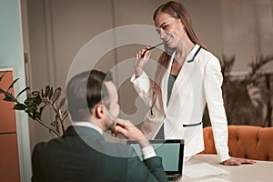 Young woman sitting on the table and sexually looking at young man. Two young and attractive colleagues man and woman