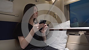 Young woman is sitting in a train carriage near the window. She communicates on her smartphone and looks out the window