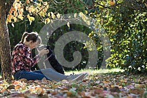 Young woman sitting under a coulourful autum tree lovingly petting her black dog