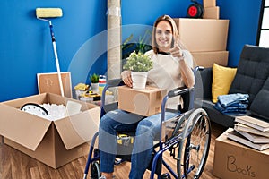 Young woman sitting on wheelchair moving to a new home pointing fingers to camera with happy and funny face