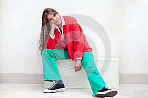 young woman sitting on white box wearing red jacket and green trousers