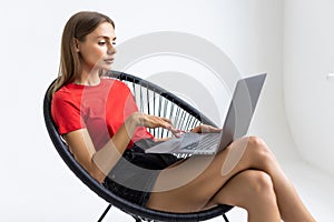 Young woman sitting on a white chair with laptop in an empty room, thinking on something