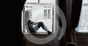 Young woman sitting on window sill and enjoying old town view. Silhouette of relaxing Girl on fresh air at home