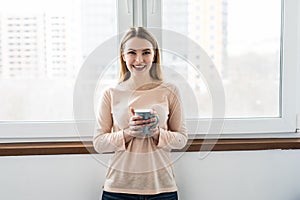 Young woman sitting on the windowsill and looking out the window with cup of coffee in the morning