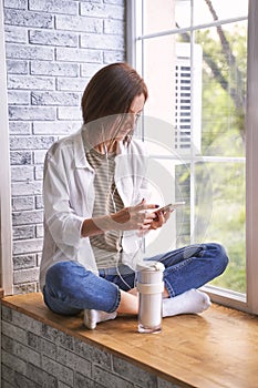 Young woman sitting at windowsill. Working on phone. Online study concept