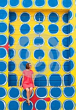 A young woman sitting on a yellow wall with a pattern of blue-white dots