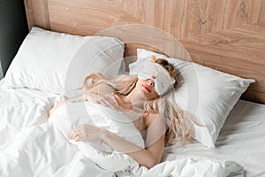 Young woman sleep on comfortable bed in a mask for sleeping. Blindfold on eye. Morning in hotel room. White pillow and