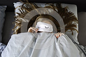 Young woman is sleeping in bed with sleep mask and peeking from underneath the sleeping mask