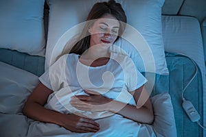 Young woman sleeping on electric heating pad in bed at night, top view