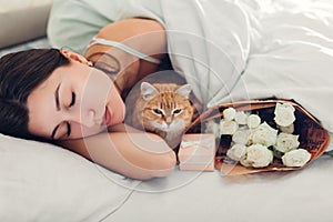 Young woman sleeping near bouquet of roses and gift box in bed with cat. Present left by boyfriend for Women`s day