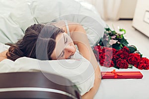 Young woman sleeping near bouquet of roses and gift box in bed. Present left by boyfriend for Valentines day