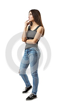 Young woman in sleeveless gray top and blue jeans standing in half turn, deep in thought, with fingers rubbing her chin