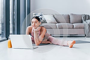 Young woman with slim body shape in sportswear sits on mat with orange juice and laptop indoors at home