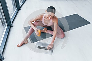 Young woman with slim body shape in sportswear sits on mat with orange juice and laptop indoors at home