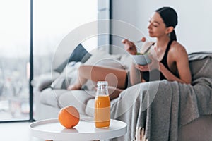 Young woman with slim body shape in sportswear sits on sofa and eats healthy diet food indoors at home