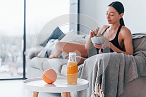 Young woman with slim body shape in sportswear sits on sofa and eats healthy diet food indoors at home